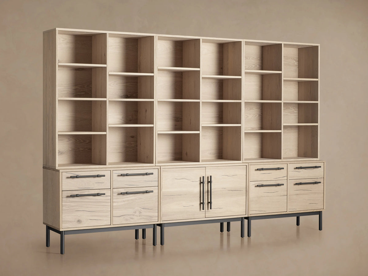 Sullivan Triple Bookcase in natural oak finish, showcasing adjustable shelves and iron-handled drawers. Placed on a wooden floor.