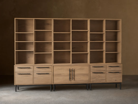 Sullivan Triple Bookcase in natural oak finish, showcasing adjustable shelves and iron-handled drawers. Placed on a wooden floor.