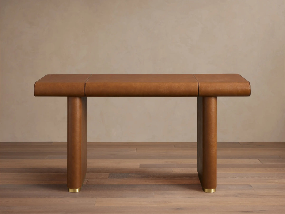 Tullson Leather Desk with smooth brown leather finish, four cylindrical legs, and gold accents at the base, on wooden floor.