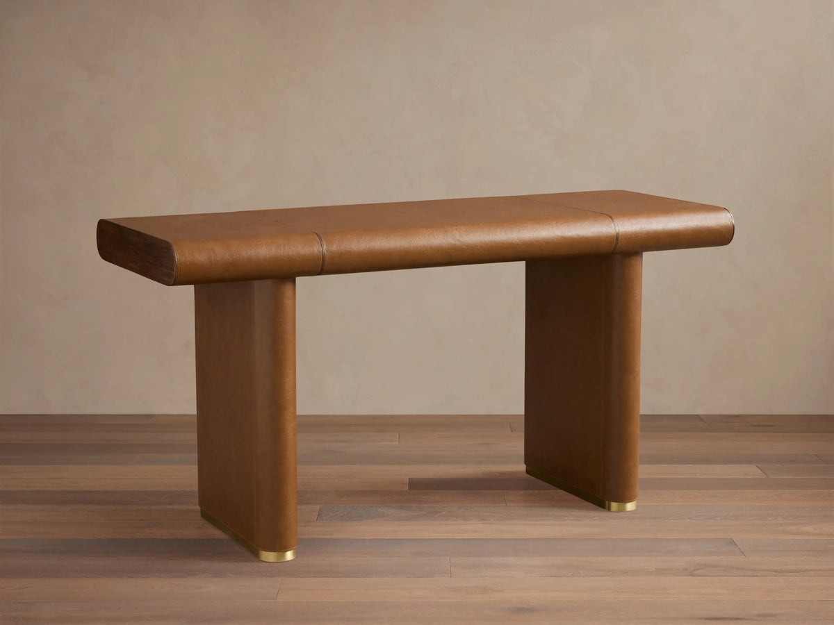 Tullson Leather Desk with smooth brown leather finish, four cylindrical legs, and gold accents at the base, on wooden floor.