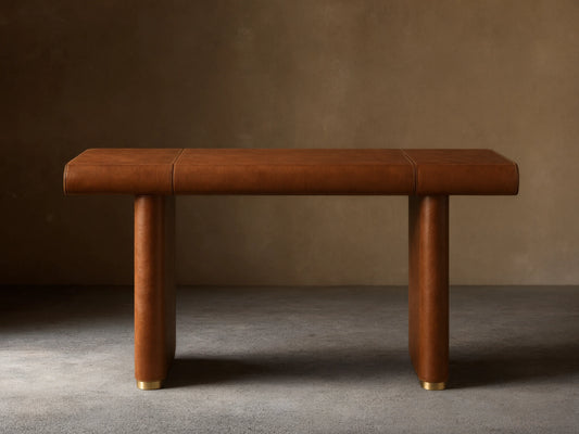 Tullson Leather Desk with smooth brown leather finish, four cylindrical legs, and gold accents at the base, on wooden floor.
