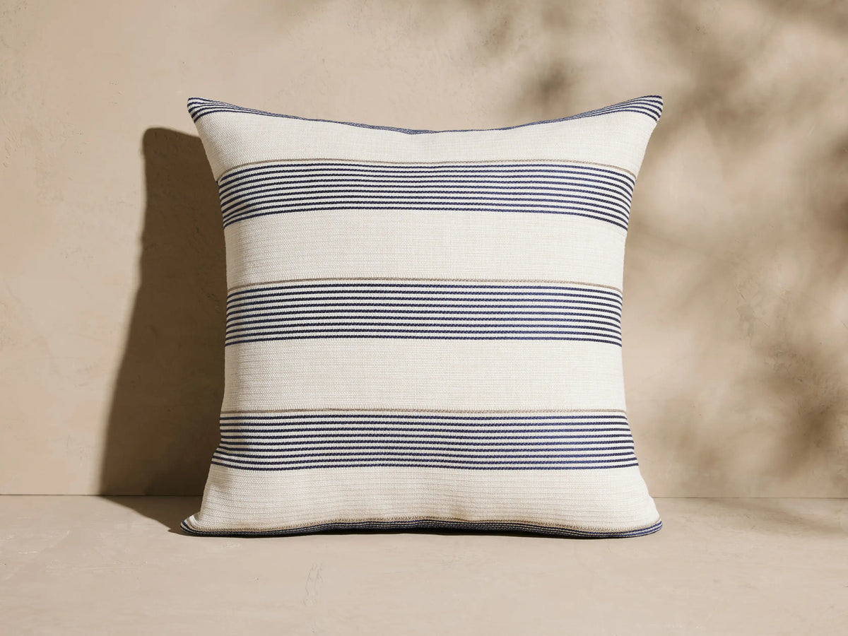 Dawson Outdoor Pillow, cream square with horizontal navy and tan woven stripes, upright against a beige textured wall.