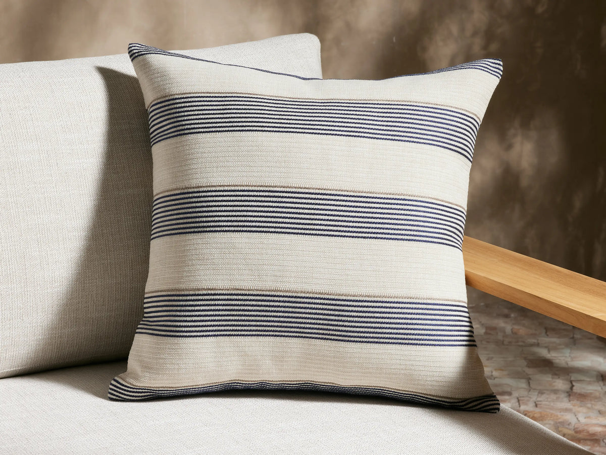 Dawson Outdoor Pillow, cream square with horizontal navy and tan woven stripes, upright against a beige textured wall.