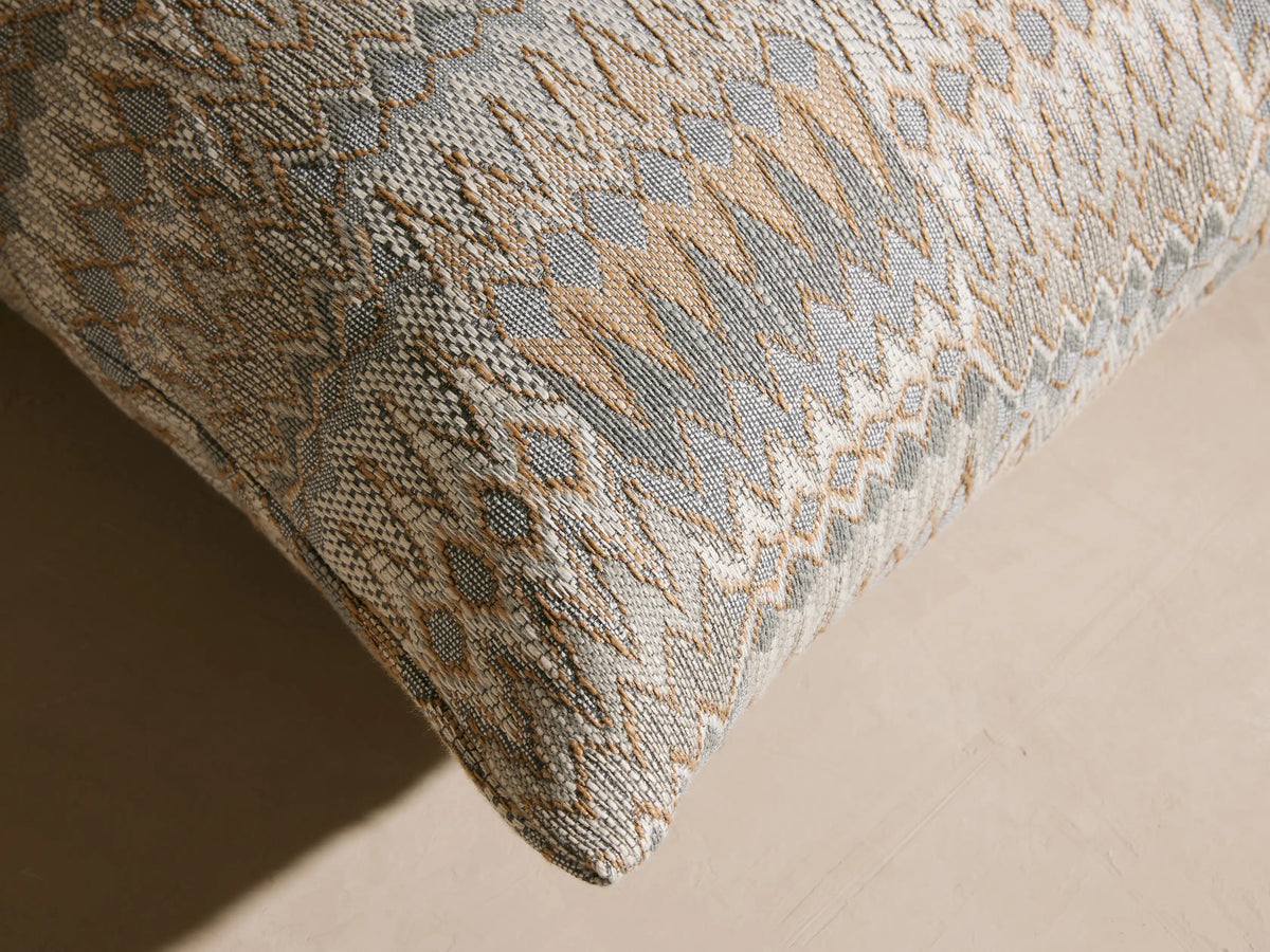 Square Naya Outdoor Pillow with intricate gray, beige, and white jacquard pattern on a light outdoor sofa in a patio setting.