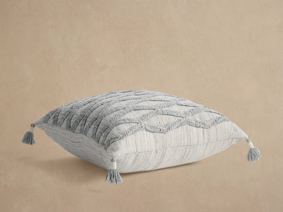 Mesa Outdoor Floor Pillow, light gray woven square with subtle vertical stripes & gray tassels on each corner, flat on tan.