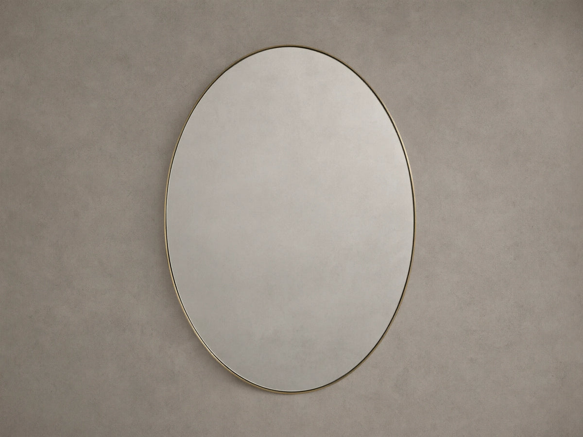Oval wall mirror with a thin brass frame, mounted on a neutral beige wall, reflecting soft ambient light.