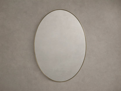 Oval wall mirror with a thin brass frame, mounted on a neutral beige wall, reflecting soft ambient light.