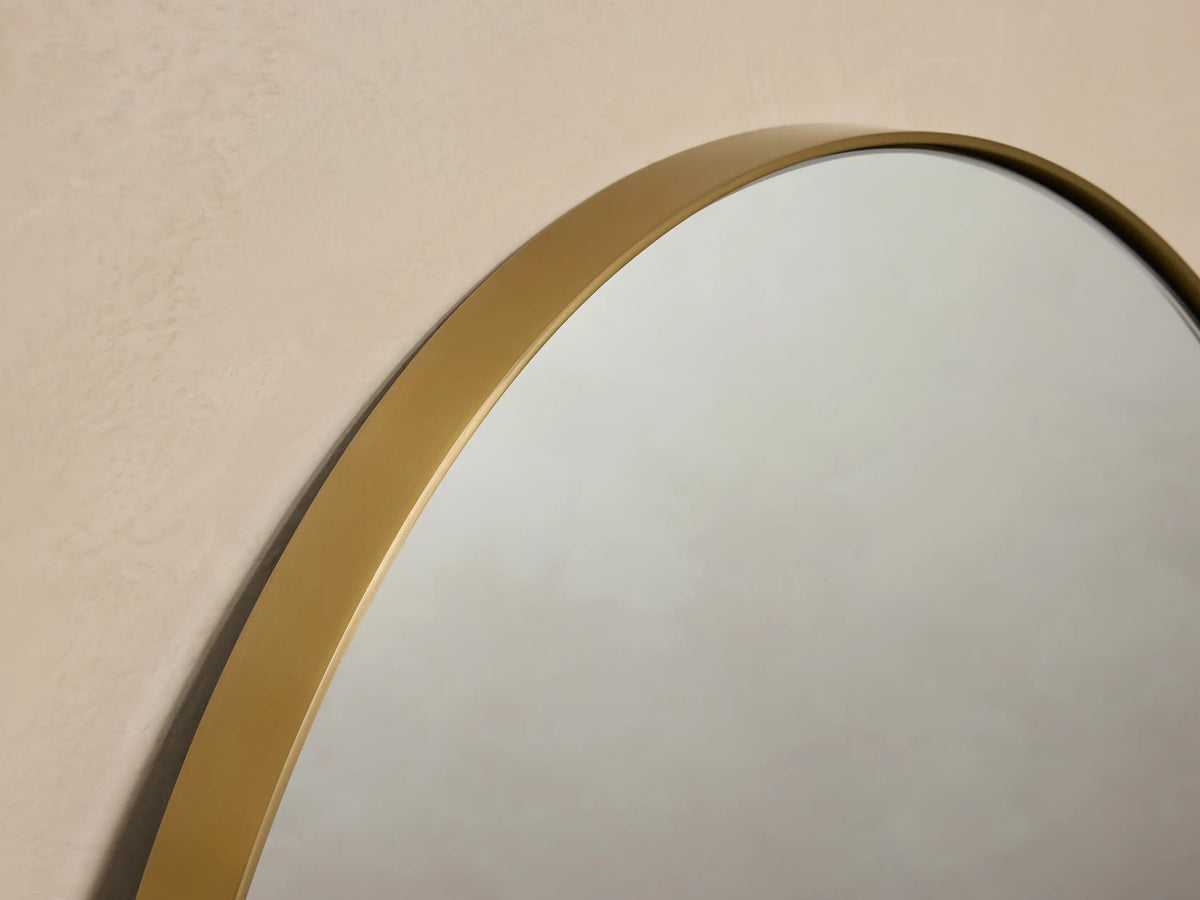 Oval wall mirror with a thin brass frame, mounted on a neutral beige wall, reflecting soft ambient light.