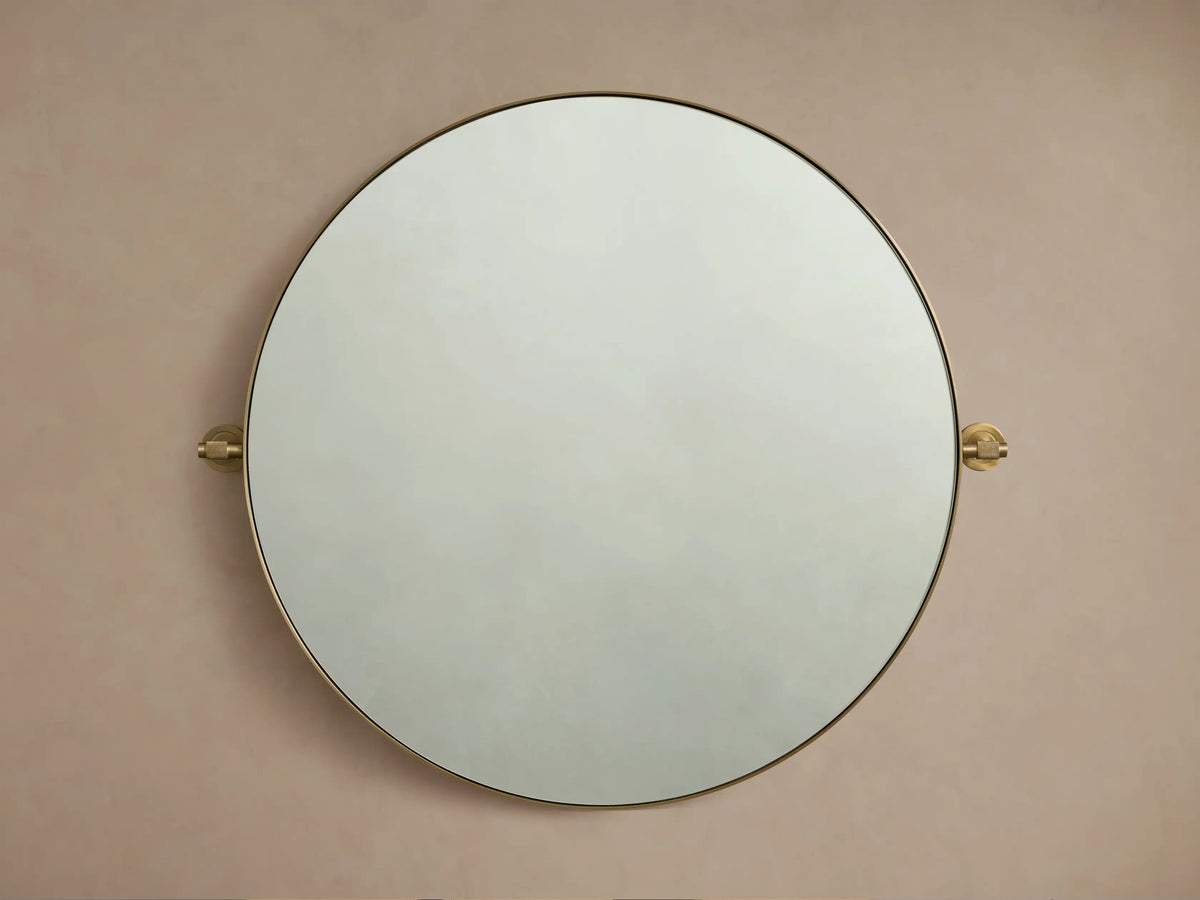 Round wall-mounted mirror with a thin brass frame, viewed frontally against a beige wall, with decorative brass mounts on sides.