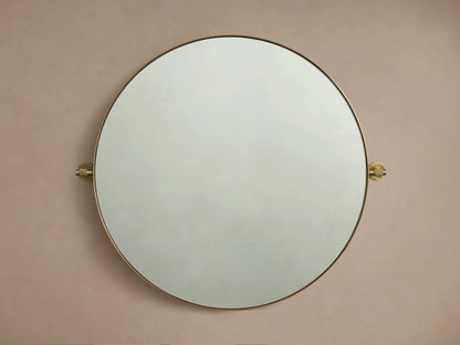 Round wall-mounted mirror with a thin brass frame, viewed frontally against a beige wall, with decorative brass mounts on sides.
