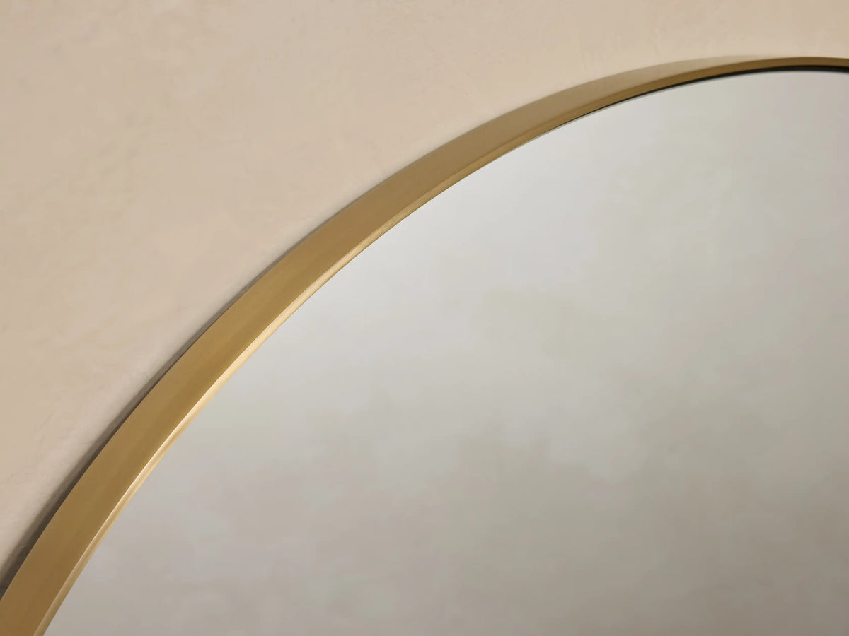 Round wall-mounted mirror with a thin brass frame, viewed frontally against a beige wall, with decorative brass mounts on sides.