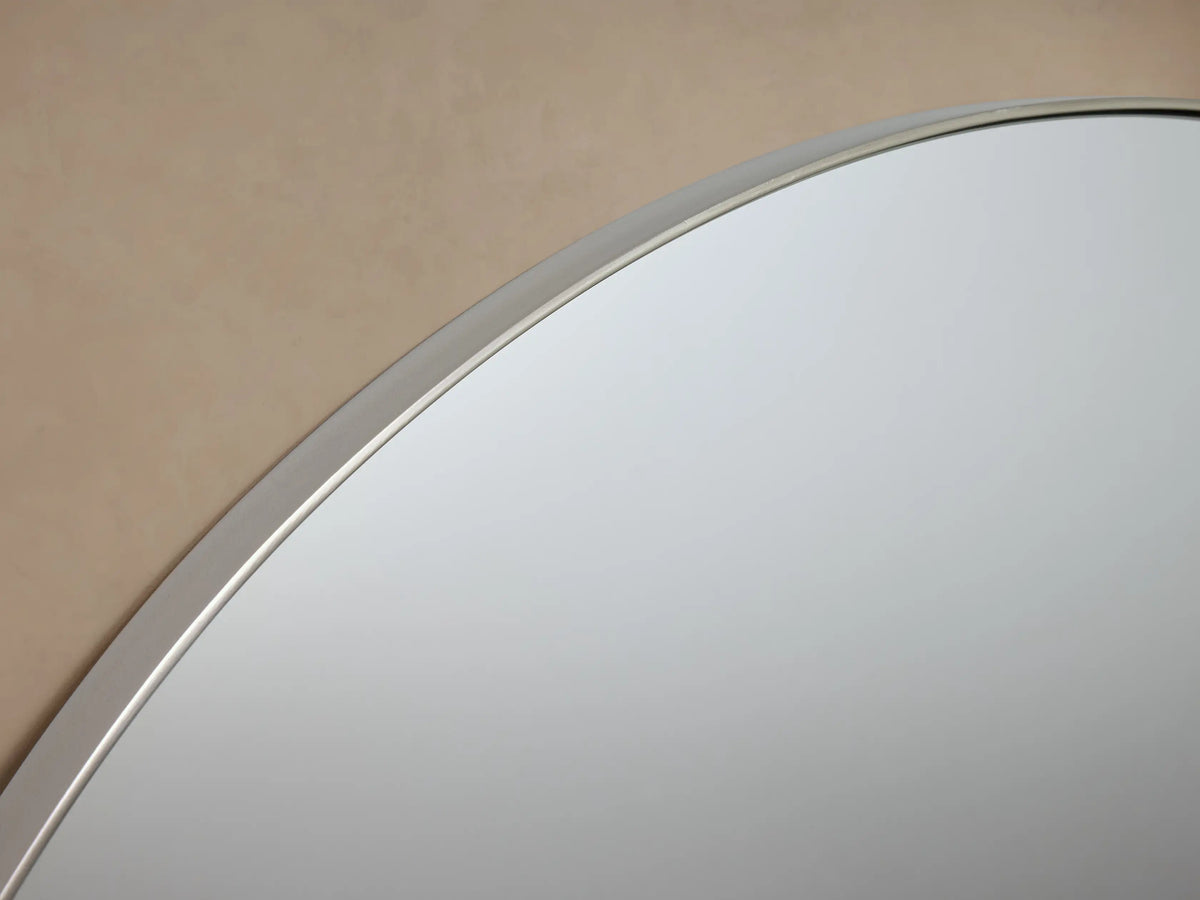 Round wall-mounted mirror with a thin metal frame, set against a beige background, featuring decorative knobs on each side.