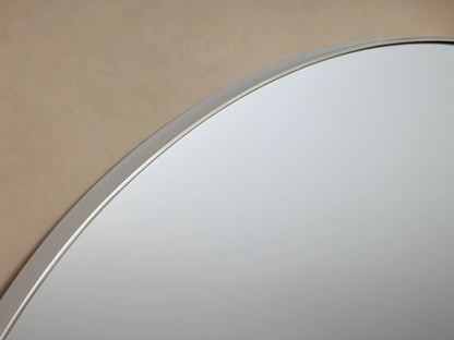 Round wall-mounted mirror with a thin metal frame, set against a beige background, featuring decorative knobs on each side.