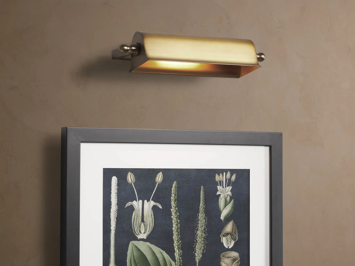 Antiqued brass Tremont Picture Light Sconce, horizontally mounted on a light beige textured wall, with finials.