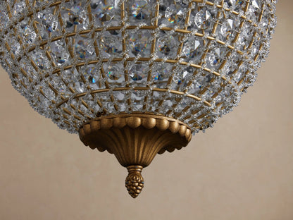 Poppy Chandelier, a spherical fixture covered in clear multifaceted crystals, hanging from an antiqued brass chain against a beige wall.