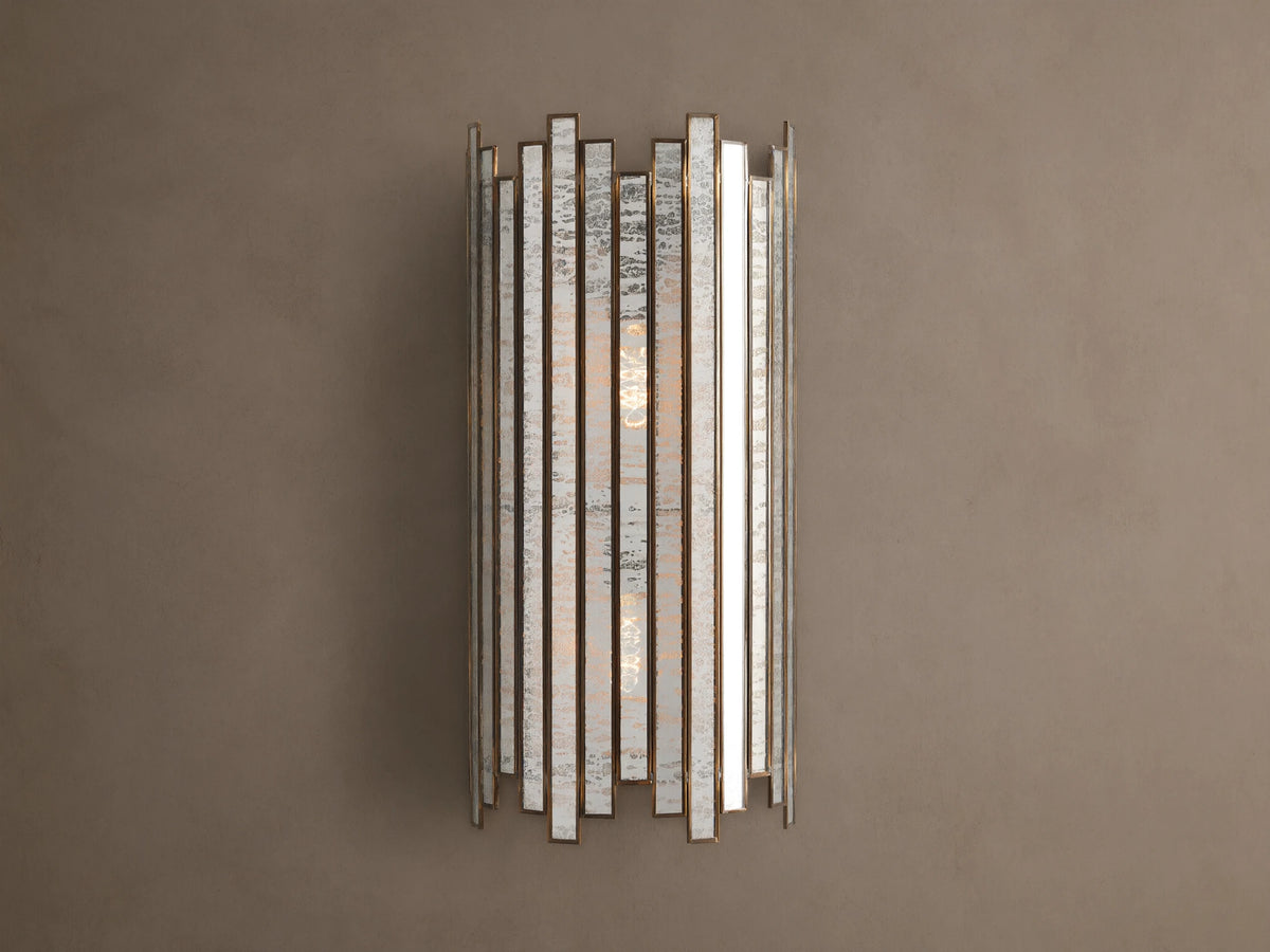 Laila Sconce: vertical antique copper frame with rectangular antiqued mirrored-glass panels, glowing softly against a beige wall.