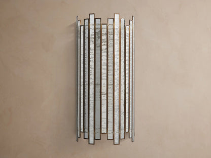 Laila Sconce: vertical antique copper frame with rectangular antiqued mirrored-glass panels, glowing softly against a beige wall.