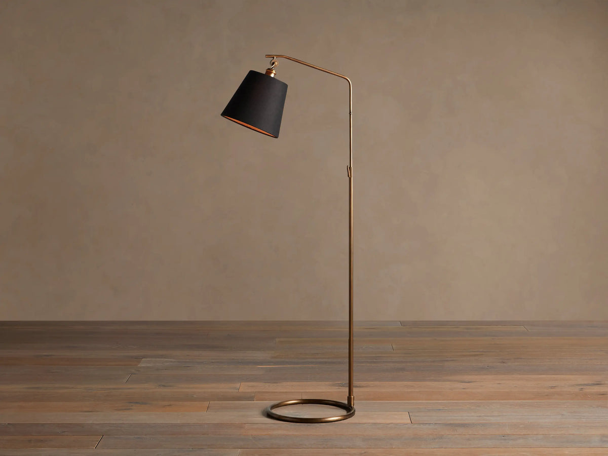 Kellen Antiqued Brass Floor Lamp, slender frame, black fabric shade with warm interior, round base on wooden floor.
