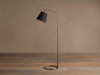 Kellen Antiqued Brass Floor Lamp, slender frame, black fabric shade with warm interior, round base on wooden floor.
