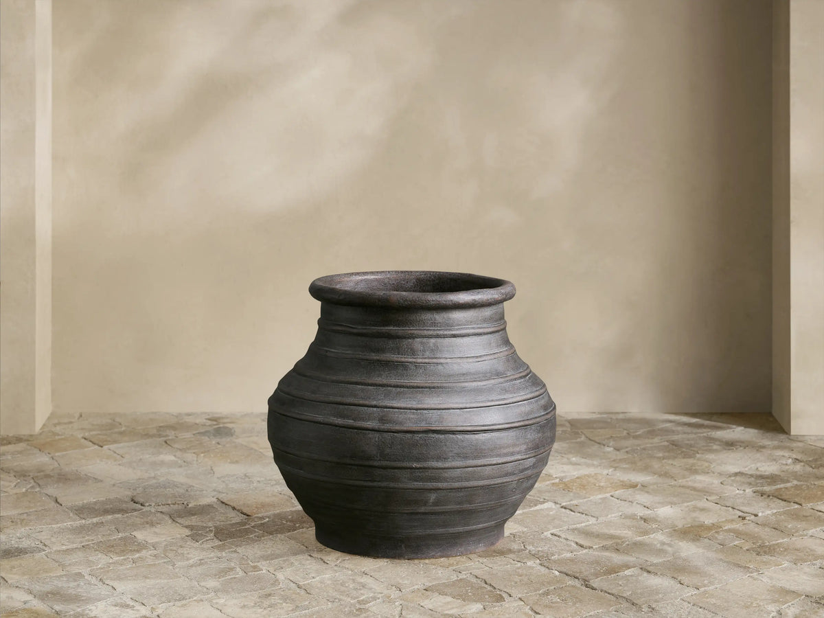 Artem Outdoor Planter: dark weathered gray, urn-shaped with horizontal ridges, on a light stone patio against a pale wall.