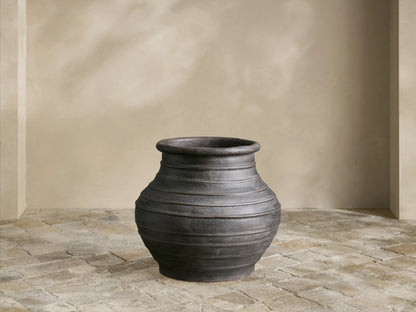 Artem Outdoor Planter: dark weathered gray, urn-shaped with horizontal ridges, on a light stone patio against a pale wall.