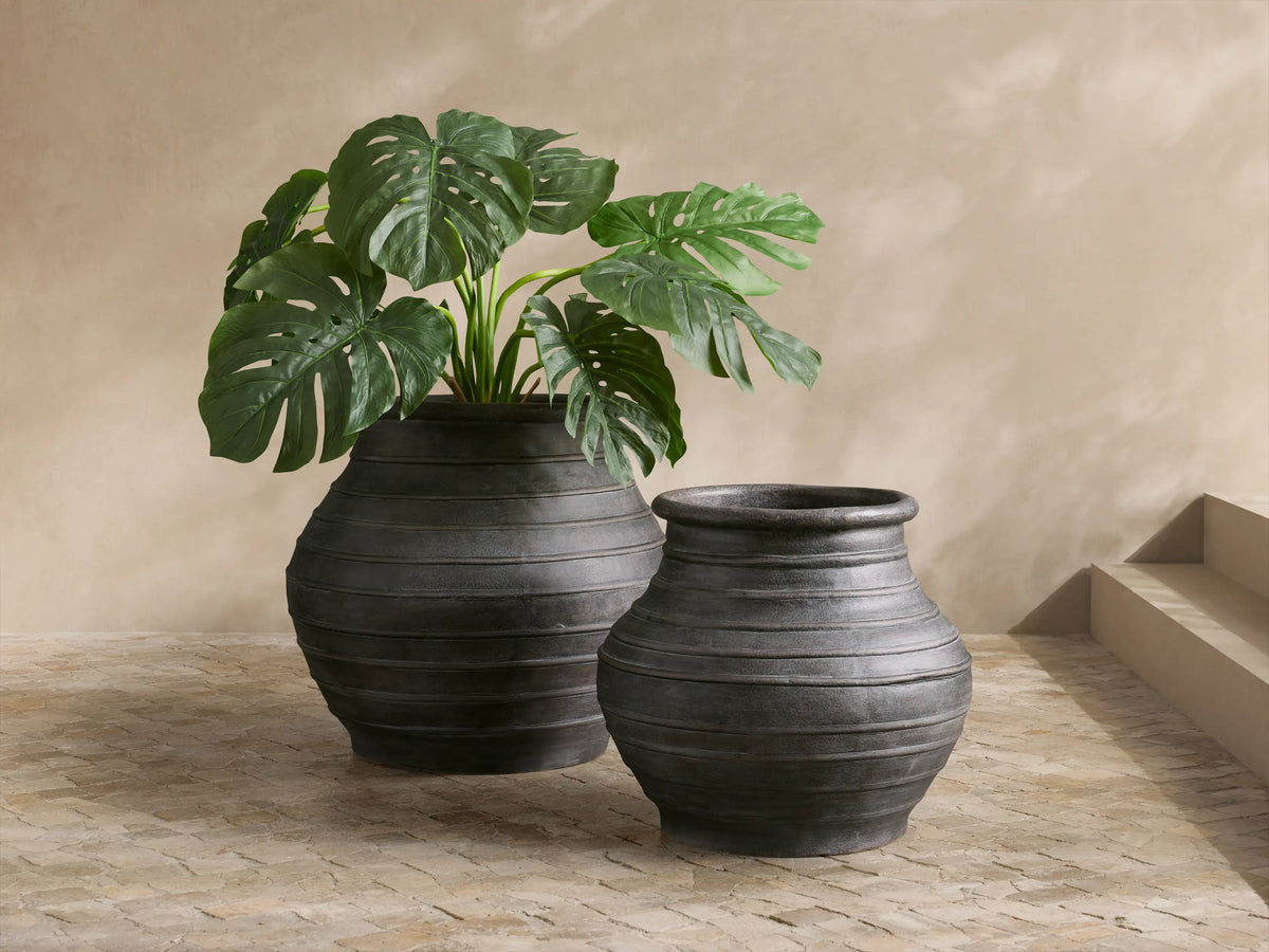 Dark, textured Artem Outdoor Planter with horizontal ridges, holding a large green plant, on a light-colored stone patio.