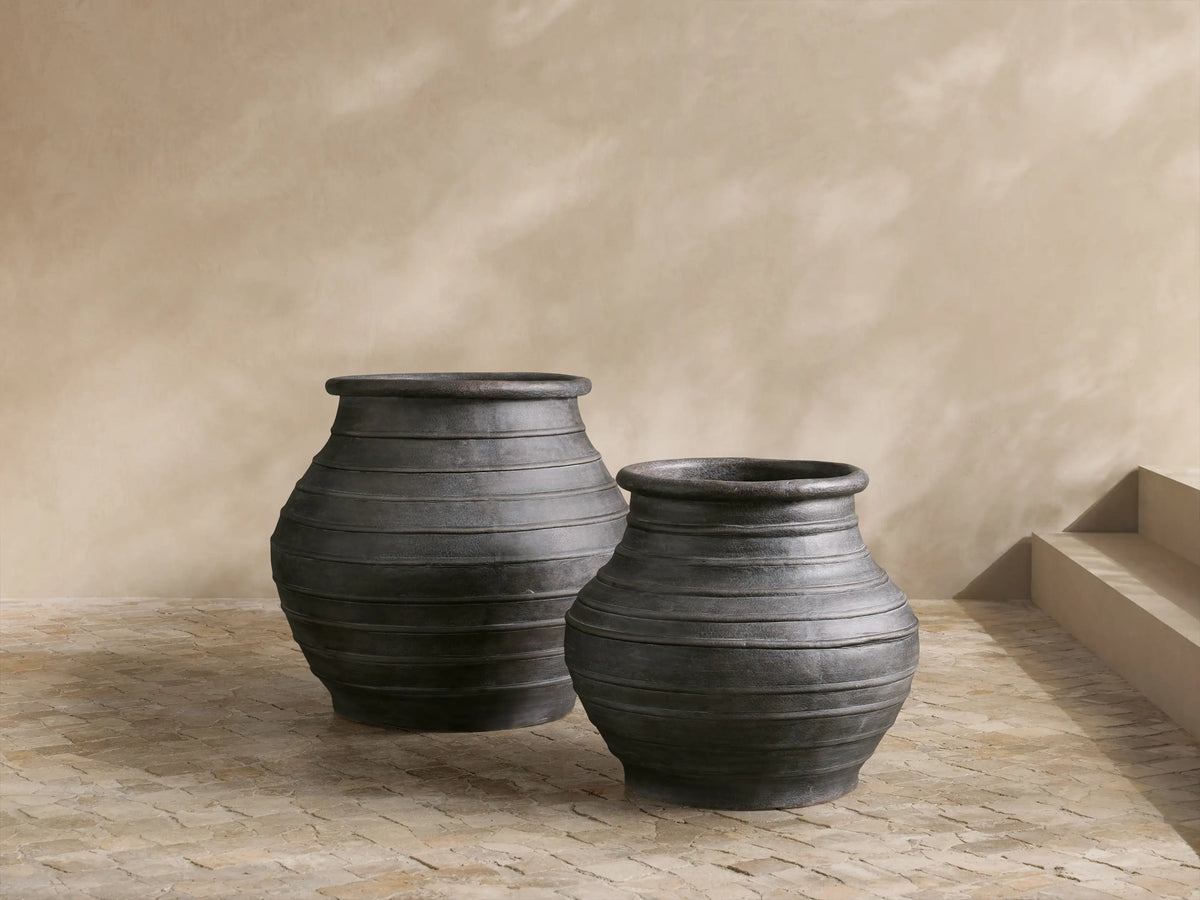 Two dark, weathered, ribbed Artem Outdoor Planters; one large with a green plant, one smaller, on a patio.