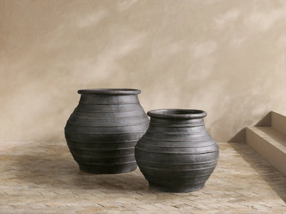 Two dark, weathered, ribbed Artem Outdoor Planters; one large with a green plant, one smaller, on a patio.