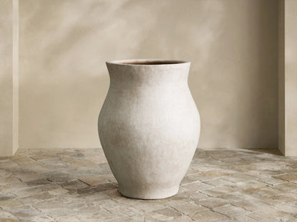 Large, curved Gable Outdoor Planter, light gray with weathered finish, sitting on textured stone pavers by a beige wall.