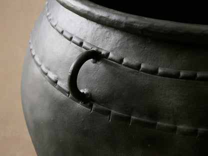 Ilya Outdoor Planter: large, dark charcoal aged metal planter with two ring handles and decorative stitching, on stone floor.