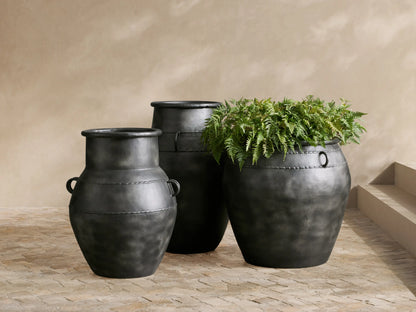 Urn-shaped Ilya Outdoor Planter, dark charcoal aged aluminum, holding a green olive tree on a stone patio.