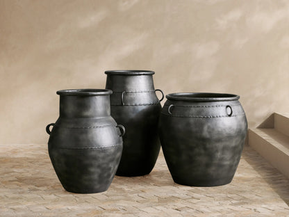 Three dark charcoal Ilya Outdoor Planters on a patio. Varied shapes, aged metal patina. Largest holds ferns; one has side handles.