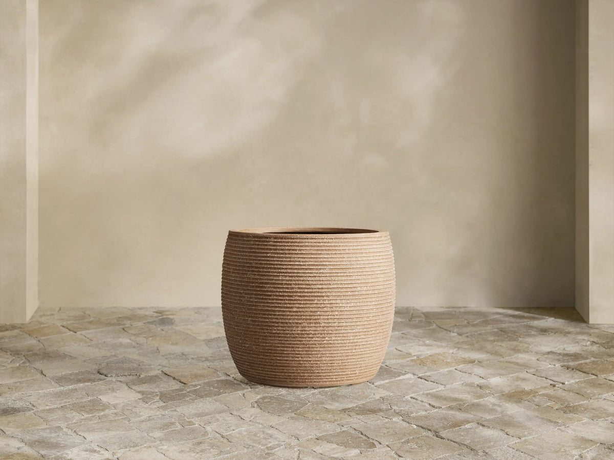 Medium brown ribbed Ojai Outdoor Planter, filled with bright green fern, sits on a stone patio against a light wall.