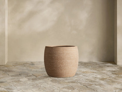 Medium brown ribbed Ojai Outdoor Planter, filled with bright green fern, sits on a stone patio against a light wall.