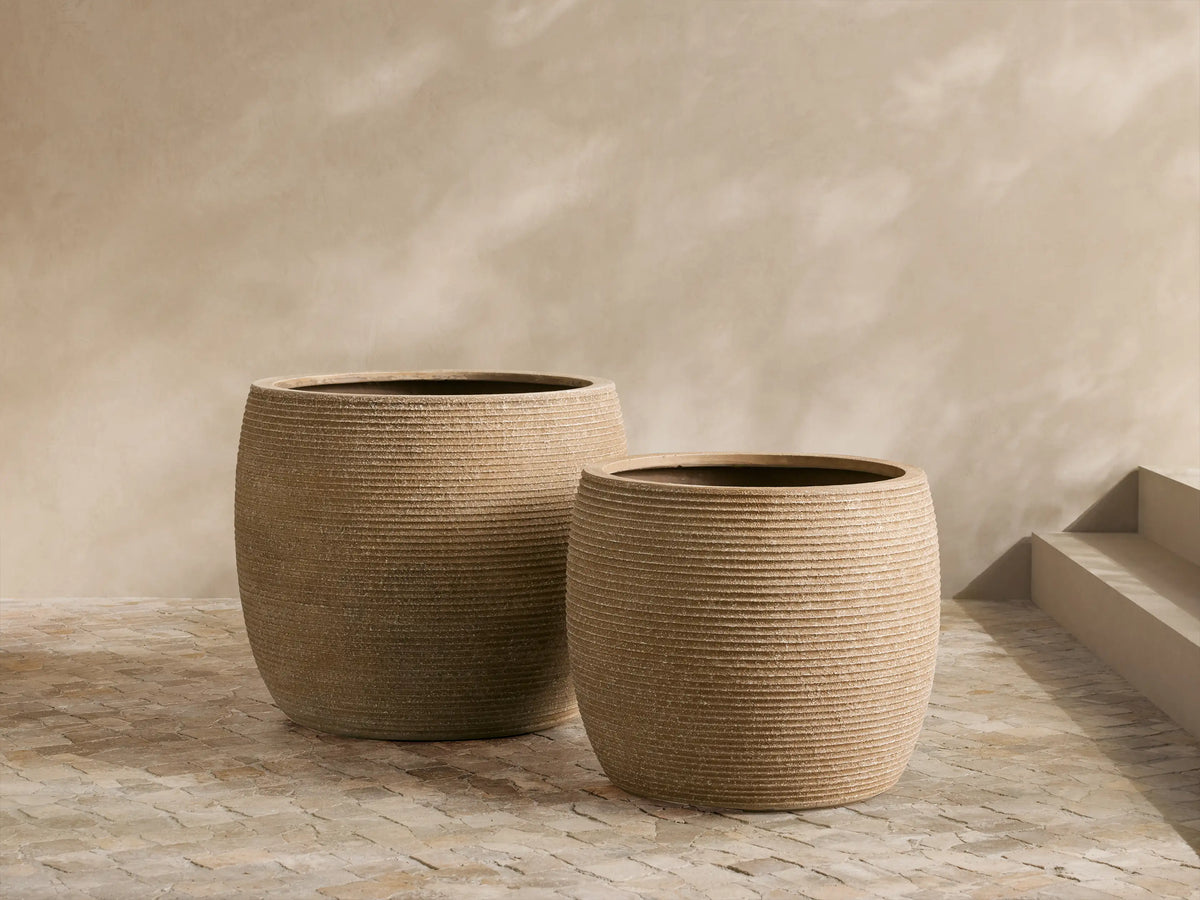 Ojai Ribbed Outdoor Planter, light brown fiberclay, cylindrical with horizontal ribs, on stone patio against a tan wall.