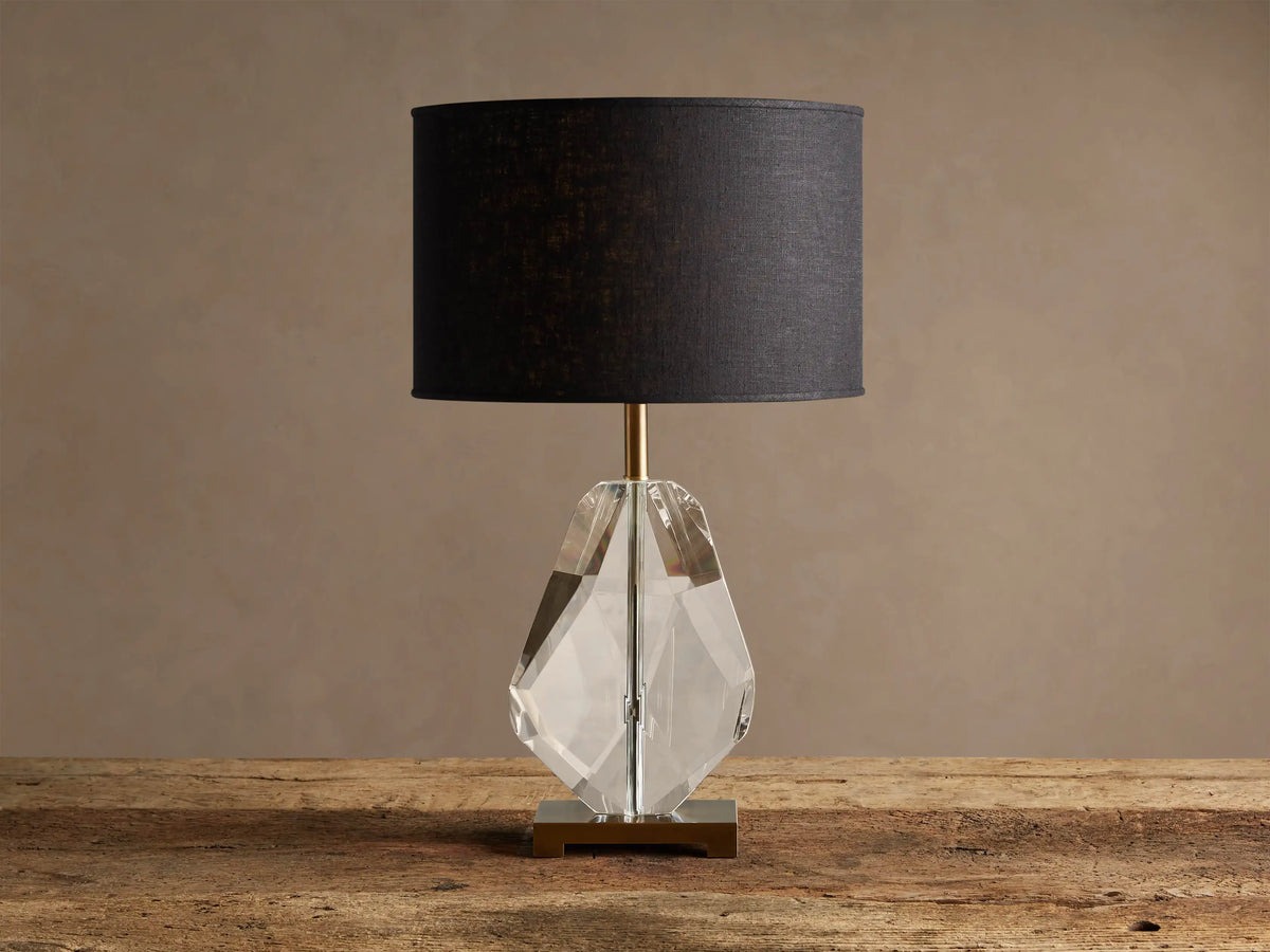 Lola Teardrop Table Lamp with black drum shade, clear faceted crystal base, and brass accents, on a wooden table.