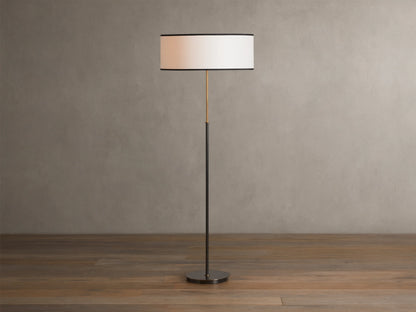 Daphne Floor Lamp with a slim bronze-finished stand, round white linen shade with black trim, wooden floor background.