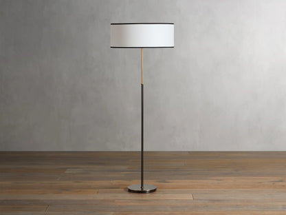 Daphne Floor Lamp with a slim bronze-finished stand, round white linen shade with black trim, wooden floor background.