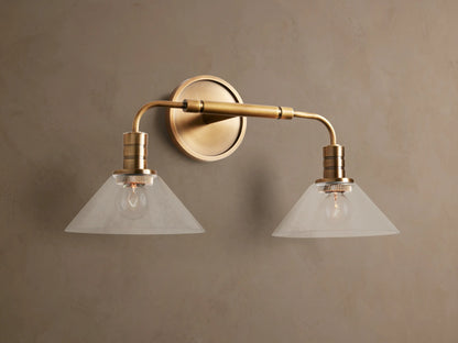 Theron Double Sconce with a brass finish, featuring two milky glass cone shades, horizontally mounted on a taupe wall.