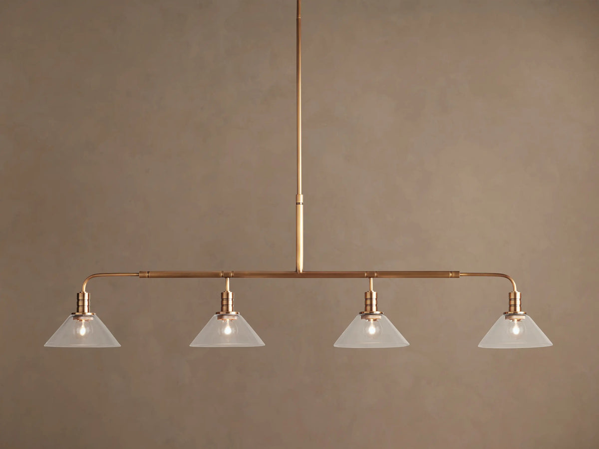 Theron Linear Chandelier: horizontal brass bar, 4 lit bulbs inside milk glass cone shades, hanging against brown wall.