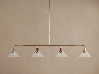 Theron Linear Chandelier: horizontal brass bar, 4 lit bulbs inside milk glass cone shades, hanging against brown wall.