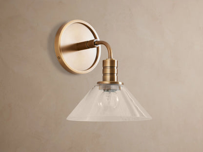 Theron Sconce, a brass wall-mounted light with a round backplate, curved arm, and clear glass cone shade, lit, on a taupe wall.