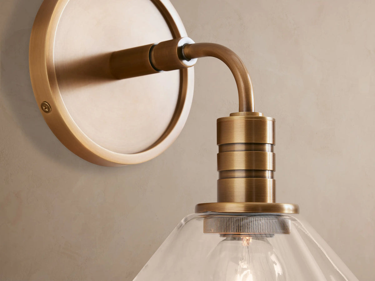 Theron Sconce: Brass-finished steel wall sconce with round backplate, curved arm, and clear glass cone shade on a tan wall.