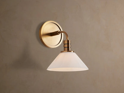 Theron Sconce with brass finish and white milk glass cone, mounted on a textured tan wall, emitting a soft glow.