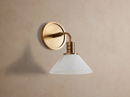 Theron Sconce with brass finish and white milk glass cone, mounted on a textured tan wall, emitting a soft glow.
