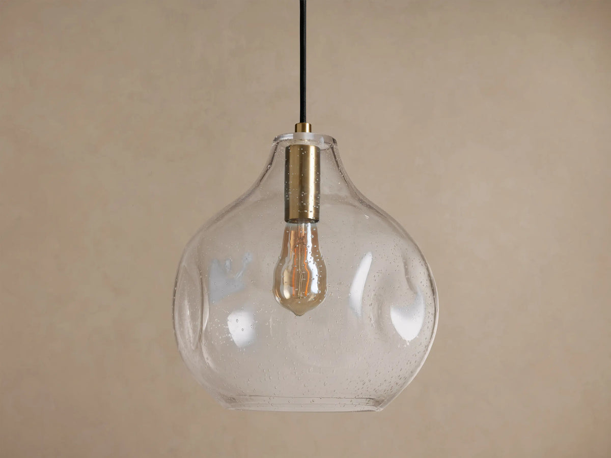 Organic 12" Pendant. Clear, mouth-blown seeded glass light fixture with a brass neck & visible filament bulb, against a beige wall.