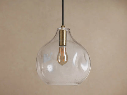 Organic 12" Pendant. Clear, mouth-blown seeded glass light fixture with a brass neck & visible filament bulb, against a beige wall.