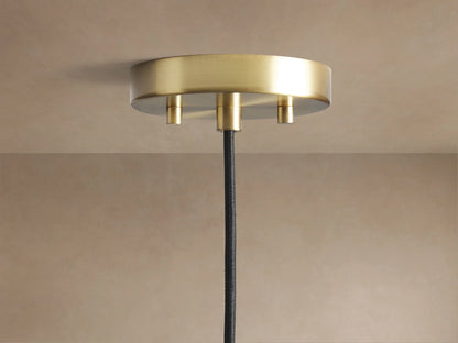 Organic 12" Pendant's round, brushed brass ceiling mount with a black fabric cord hanging from a light beige ceiling.