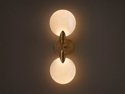Lyla Double Sconce with two glowing spherical glass shades on a brass fixture, mounted vertically on a neutral wall.