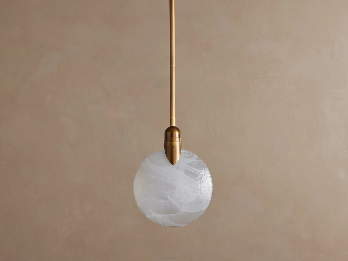 Lyla Single Pendant, a brass-plated fixture with a glowing, spherical hand-blown glass shade featuring white swirls.